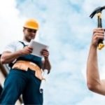 HVAC technician in safety gear using a tablet, while another worker holds a hammer, outdoors with a clear sky, emphasizing safety and professionalism in home service.