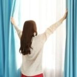 Woman opening blue curtains to let in natural light, representing home comfort and energy efficiency in relation to HVAC services.
