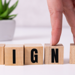 Hand arranging wooden blocks spelling "SIGN" with a small potted plant in the background, emphasizing home maintenance and property management themes.