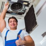 HVAC technician smiling and giving a thumbs up while holding an air conditioning unit, representing professional AC services and maintenance.