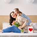 Woman and young girl in cozy indoor setting with bouquet of flowers and heart box, symbolizing affection.