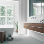 Modern bathroom with wooden vanity, large mirror, and bathtub, featuring natural light from windows and greenery.