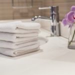 Stack of white towels on a bathroom countertop beside a faucet and a vase with a purple flower, emphasizing guest bathroom readiness for visitors.