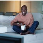 Person sitting on a couch enjoying popcorn while watching a movie, creating a cozy atmosphere for a horror movie night.