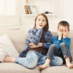 Children watching a scary movie on a cozy couch, with one child covering their eyes and the other looking surprised, highlighting the importance of a warm and inviting space for movie nights.