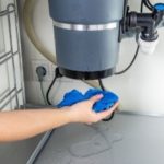 Hand holding a blue sponge near a garbage disposal unit under a kitchen sink, illustrating maintenance tips for addressing common garbage disposal issues.