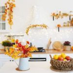 Brightly decorated kitchen with autumn foliage, fruits in a basket, and warm lighting, creating a cozy atmosphere for home gatherings.