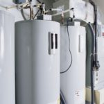Water heaters mounted on a wall with gray insulation, pipes, and valves at Carmine's Plumbing, Heating & Air Conditioning.