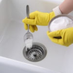 Person in yellow gloves adding baking soda to a kitchen sink drain, emphasizing home plumbing maintenance and cleaning solutions.