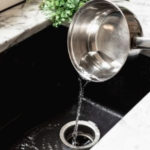 Stainless steel pot pouring water into a kitchen sink drain, illustrating common plumbing issues and the need for professional services.