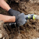 A black-gloved hand grips a leaking pipe in a muddy excavation, showcasing urgent plumbing repair work by Carmine's Plumbing.