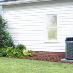 Air conditioning unit beside a white house with a well-maintained yard and shrubs, illustrating HVAC system installation and home comfort services.