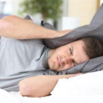 Man lying in bed with a pillow over his head, appearing distressed by noise, illustrating the discomfort caused by home maintenance issues.