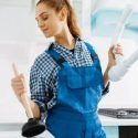 A woman in blue overalls holds a plunger, prepared for plumbing maintenance in a kitchen setting.