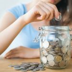 Person adding coins to a clear jar filled with coins, symbolizing savings and financial management in the context of affordable HVAC solutions.