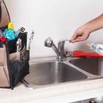 Hand adjusting faucet in kitchen sink with plumbing tools in background, illustrating plumbing maintenance services.