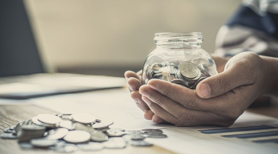 Hands holding a glass jar of coins with paperwork and a laptop, symbolizing home maintenance finance.