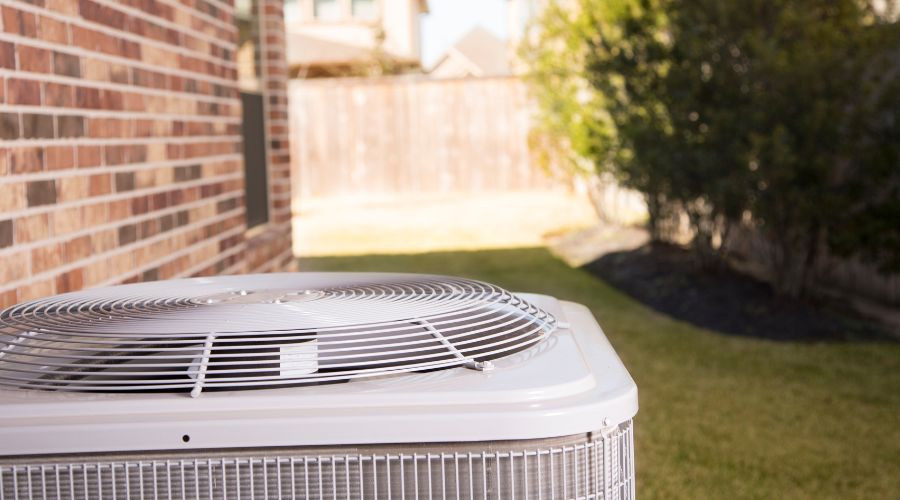 Air conditioning unit in residential backyard; features brick wall, wooden fence, and manicured lawn for home comfort.