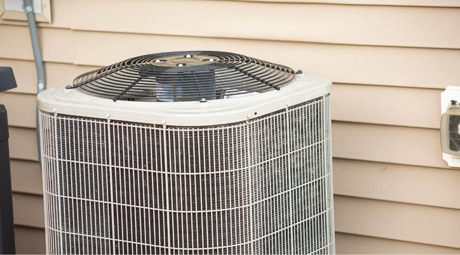 Central AC condenser with top mounted fan and protective metal cage positioned next to home exterior with electrical conduit and disconnect box visible